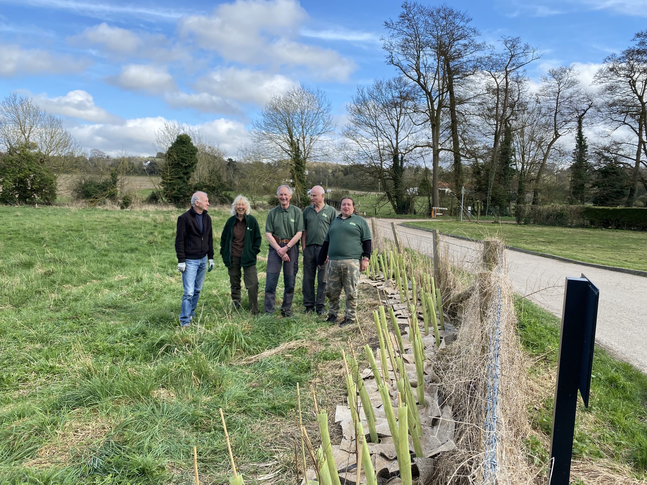 Five people dressed in green and black sanding beside a row of newly planted hedgerow trees in a green field