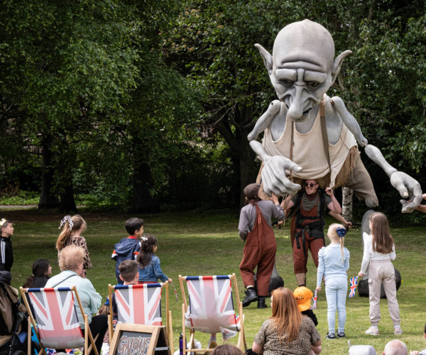 Extra large puppet being moved by three people in front of people sitting on deckchairs