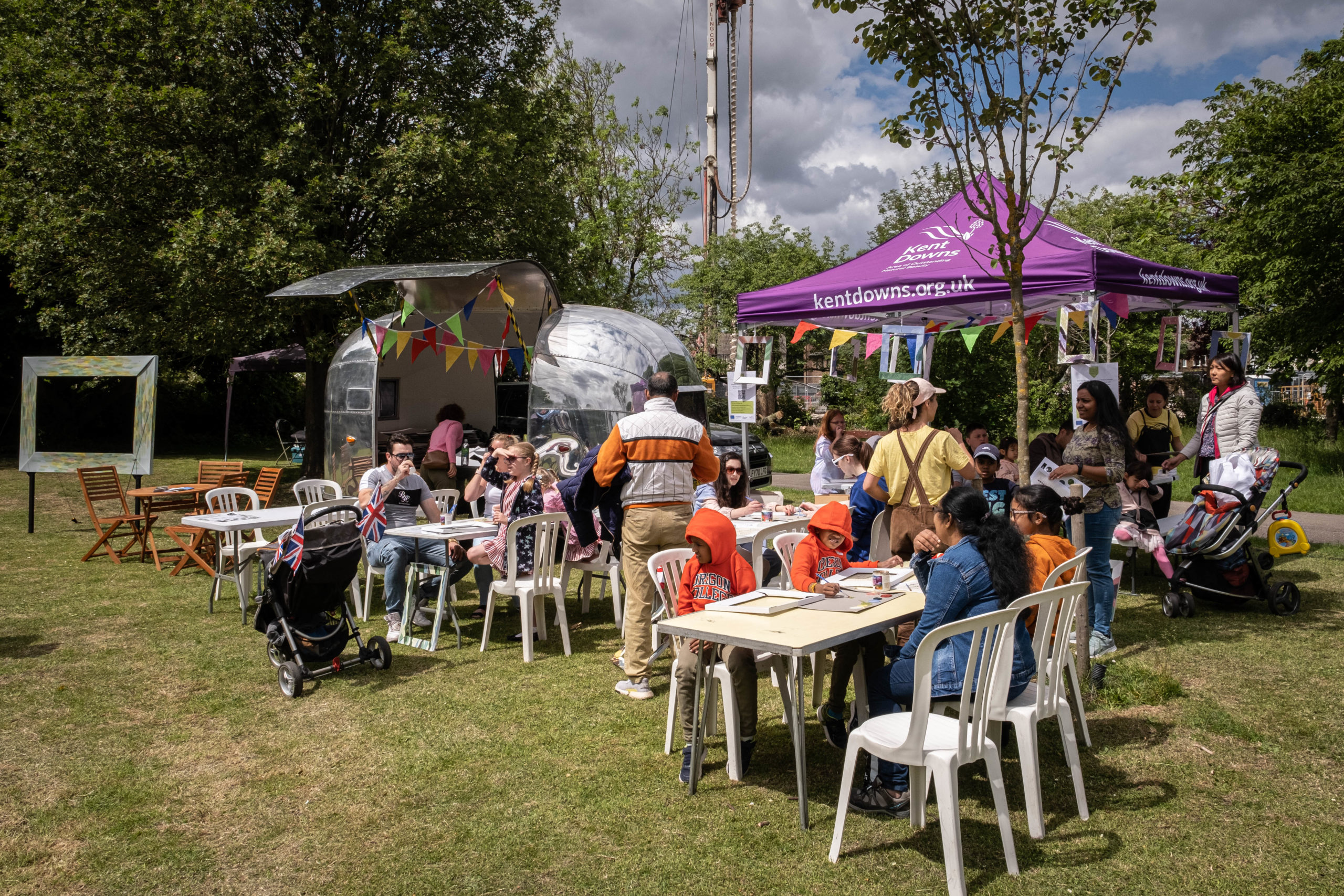 Silver airstream caravan parked next to purple gazebo with people in front sat at tables and chairs painting frames